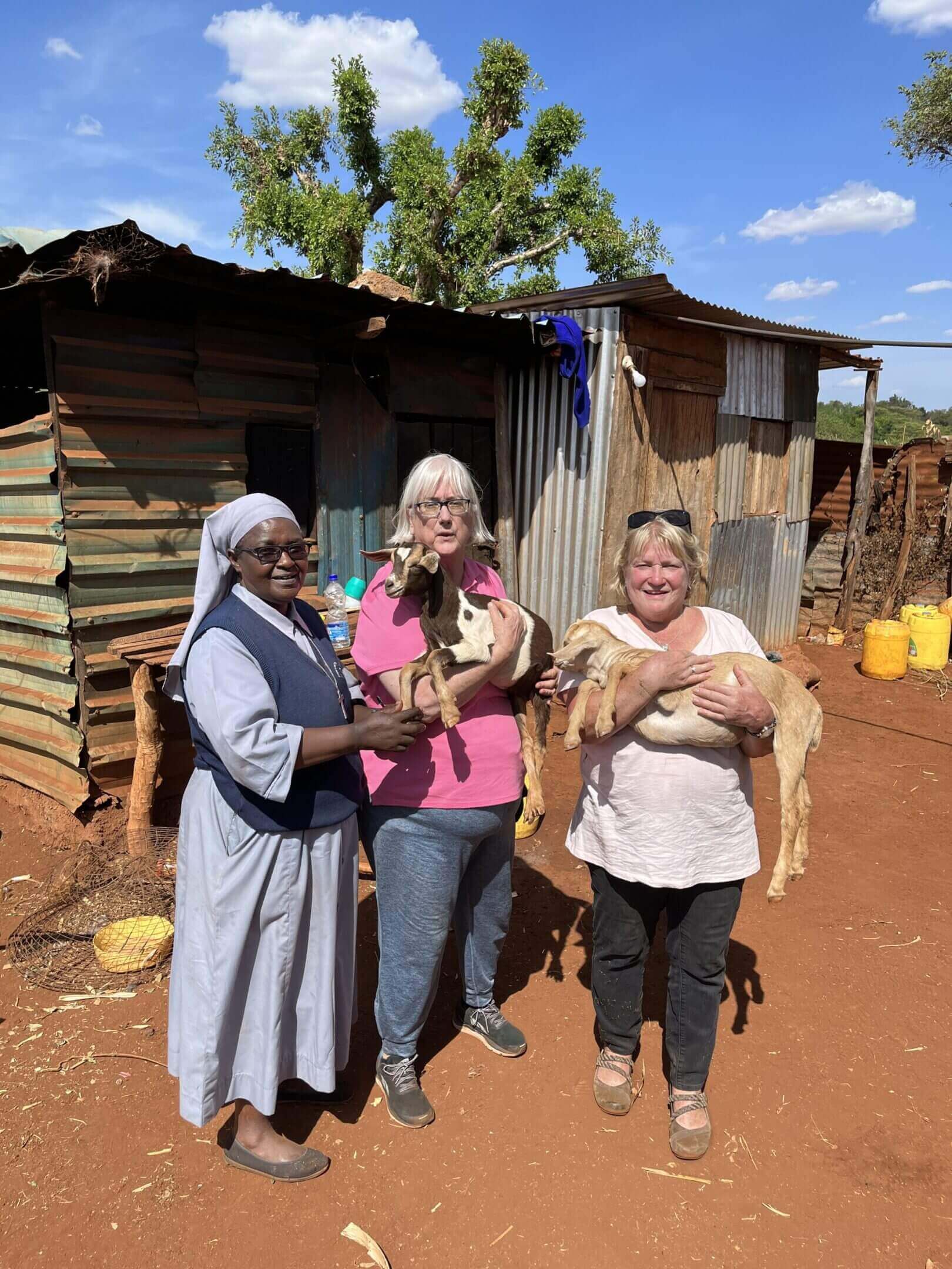 Three women holding goats near rustic building.