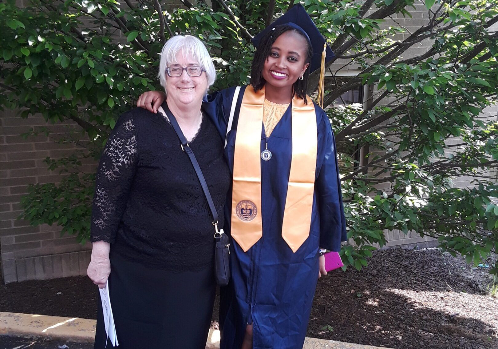 Two women posing outdoors, one in graduation attire with a gold stole.