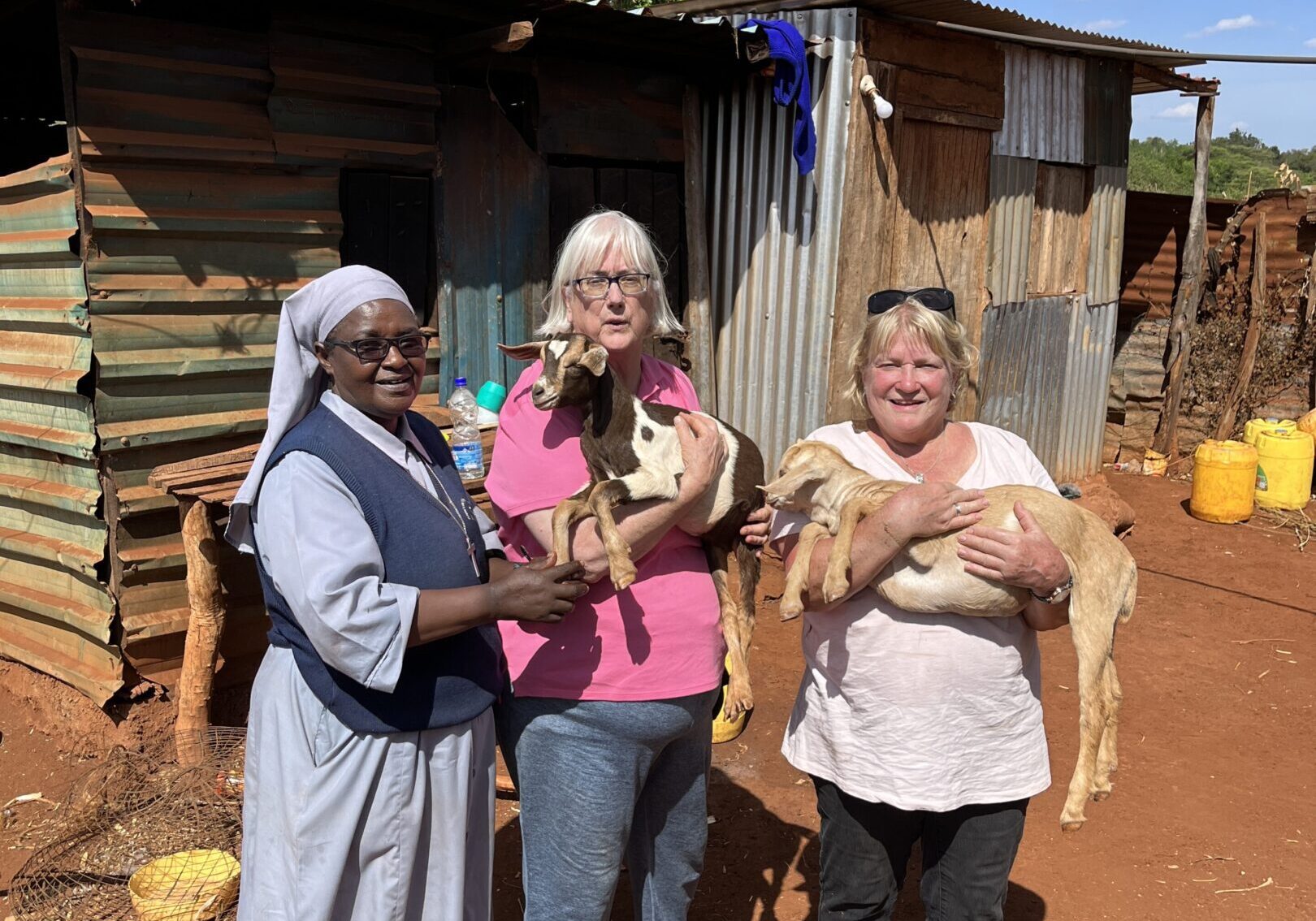 Three women smiling outdoors near rustic wooden buildings under a sunny sky.
