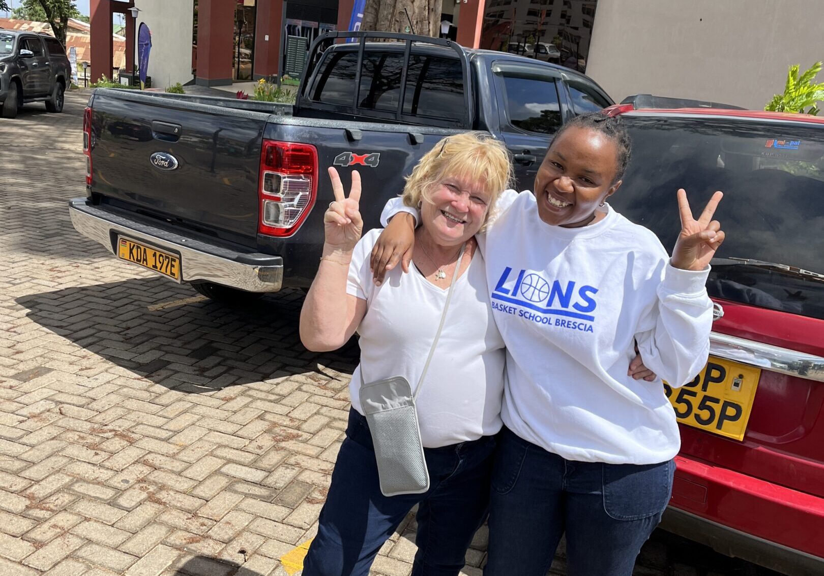 Two women smiling and posing with peace signs near parked vehicles.